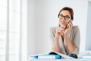 Photo of female employee has cheerful expression, talks via smartphone, hold pen, sits at desktop with papers and notepads, computer, poses against office interior, wears spectacles, elegant clothes