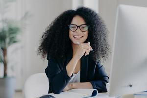 Positive international female student with crisp hair, wears transparent glasses, holds pen in hand, makes accountings, sits in front of big computer screen, dressed in black elegant outfit. photo