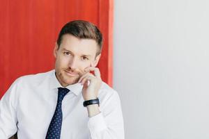 Indoor shot of self confident businessman in white shirt with tie, keeps hands near face, waits for someone, isolated over red and white background. Successful male entrepreneur poses indoor photo
