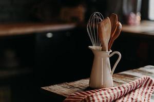 accesorios de cocina en mesa de madera. utensilios en tarro de cerámica blanca sobre fondo oscuro. estilo rústico vajilla para preparar la comida. cucharas de madera shpatula y batidor foto