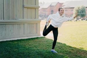 Pleased active young woman in hoodie and leggings prepares for running stratches and warms legs focused happily into distance listens music via earphones poses outdoors leads active lifestyle photo