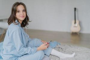 concepto de personas, descanso y estilo de vida. toma horizontal de una mujer bastante joven de cabello oscuro vestida con pijama, escucha su canción favorita a través del teléfono móvil, espacio en blanco a un lado con guitarra acústica foto