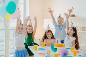 Happy birthday party. Joyful small children have fun together, raise arms and play with confetti, pose at festive table, wear party hats, being on carnival, enjoy holidays photo