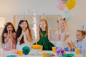 Holiday and festive event concept. Happy five small children clap hands, look at sparkle from cake, celebrate friends birthday, gather near big table, stand in decorated room with balloons photo