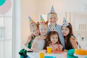 Photo of happy friends gather around table, wear party hats, embrace and look gladfully at camera, smile positively, celebrate birthday at home together, have fun