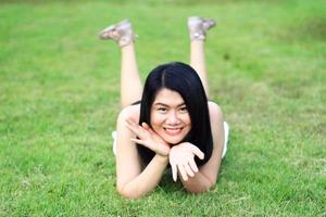 hermosa mujer asiática con vestido blanco se relaja y sonríe en un campo verde en el parque natural. joven tailandesa disfruta de vacaciones con la luz del sol en el jardín foto