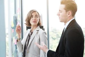 Successful businessman and businesswoman writing good ideas on sticky notes pasted on glass wall  in modern office meeting. photo