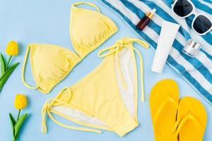 vista superior de bikini amarillo y botella de crema, sandalias, gafas de sol con accesorios de playa en el fondo azul. concepto de horario de verano. foto