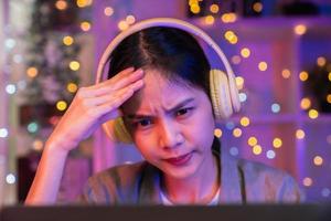 mujer asiática joven y estresante que usa auriculares y se sienta en la silla con un montón de documentos sobre la mesa y mira la pantalla de la computadora por la noche. foto