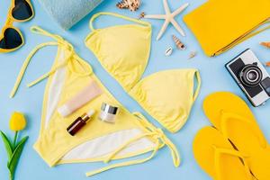vista superior de bikini amarillo y botella de crema, sandalias, gafas de sol con accesorios de playa en el fondo azul. concepto de horario de verano. foto