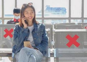 Young adult Asian female traveler sitting on public terminal seats with social distancing symbol, talking on smartphone with happy smiling face. Copy space. Safe trip after COVID-19 vaccination. photo