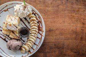 vista superior de tostadas, helado de chocolate, rodajas de plátano y crema batida en plato blanco, todo goteado con salsa de chocolate sobre fondo de mesa de madera. foto