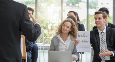 Young adult caucasian business man asking question with report in hand to speaker while making speech among the audience in corporate seminar event. Light effect. photo