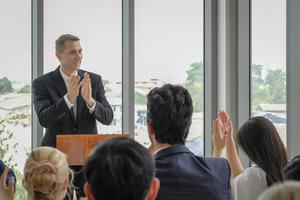 Happy caucasian businessman stand clapping hands on podium for celebration with other participants in seminar event. Selective focus. photo