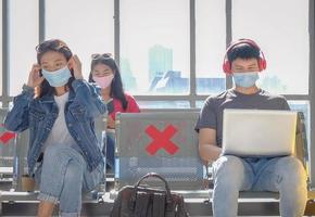 Asian young traveler group live in new normal lifestyle after COVID-19 outbreak in public area, wearing protective face mask and sitting follow distancing symbol from each other. Selective focus. photo