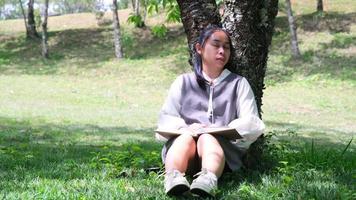 mujer asiática sentada bajo un árbol tomando una siesta mientras lee un libro en un parque. video