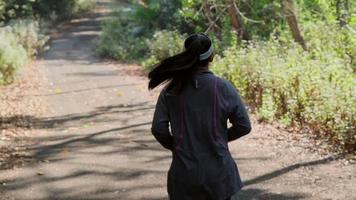 donna asiatica che pareggia sulla strada sterrata nella foresta per esercitare. donna sportiva che corre su una strada di campagna sotto alberi frondosi al mattino. video