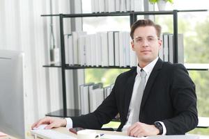 Handsome businessman in black suit wear eyeglasses and happy working with computer, notebook, laptop and smartphone on desk in modern office photo