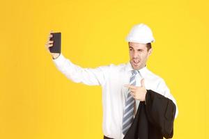 apuesto ingeniero inteligente o arquitectura lleva un sombrero de seguridad blanco con un teléfono inteligente de mano y disfruta con selfie solo aislado en fondo amarillo. copie el espacio foto