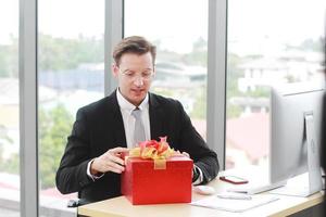 apuesto e inteligente hombre de negocios en traje negro usa gafas sorpresa y sonriendo con caja de regalo roja para el éxito en el trabajo o feliz año nuevo presente en ofiice.christmas y feliz año nuevo concepto foto