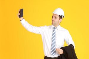 apuesto ingeniero inteligente o arquitectura lleva un sombrero de seguridad blanco con un teléfono inteligente de mano y disfruta con selfie solo aislado en fondo amarillo. copie el espacio foto