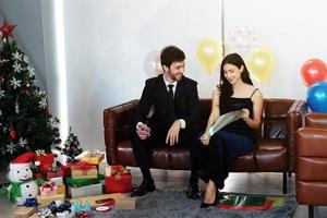 dulce pareja ama sonreír y pasar un tiempo romántico de navidad y celebrar la víspera de año nuevo en la decoración de un sofá marrón con árbol de navidad, globos coloridos y cajas de regalo en la sala de estar en casa foto