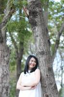 bella mujer asiática con vestido blanco se relaja y sonríe en el parque natural. chica tailandesa o chica china disfruta de vacaciones foto