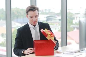 apuesto e inteligente hombre de negocios en traje negro usa gafas sorpresa y sonriendo con caja de regalo roja para el éxito en el trabajo o feliz año nuevo presente en ofiice.christmas y feliz año nuevo concepto foto