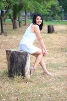 hermosa mujer asiática con vestido blanco sentada en un tocón de árbol y sonriendo en un parque natural. joven tailandesa disfruta de vacaciones con la luz del sol en el jardín. foto