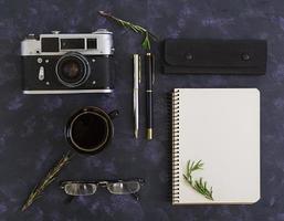 Flat lay, top view office table desk. Desk workspace with retro camera, diary, pen, glasses, case, cup of coffee, rosemary on dark background. photo