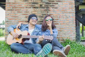 Hipster man playing guitar for his girlfriend outdoor against brick wall, enjoying together. photo