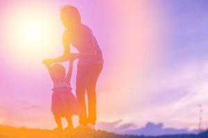 una silueta de una niña feliz los brazos de su madre amorosa para un abrazo, frente a la puesta de sol en el cielo en un día de verano. foto