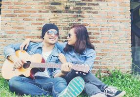 Hipster man playing guitar for his girlfriend outdoor against brick wall, enjoying together. photo