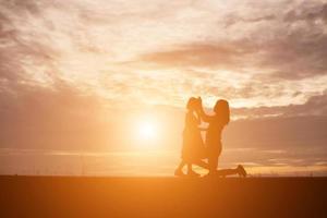 una silueta de una niña feliz los brazos de su madre amorosa para un abrazo, frente a la puesta de sol en el cielo en un día de verano. foto