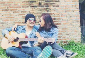 Hipster man playing guitar for his girlfriend outdoor against brick wall, enjoying together. photo