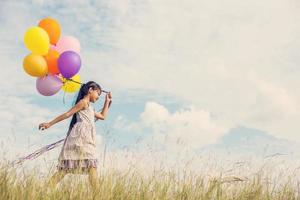 linda niña sosteniendo globos coloridos en el prado contra el cielo azul y las nubes, extendiendo las manos. foto