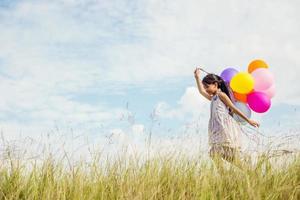 Cute little girl holding colorful balloons in the meadow against blue sky and clouds,spreading hands.