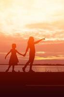 una silueta de una niña feliz los brazos de su madre amorosa para un abrazo, frente a la puesta de sol en el cielo en un día de verano. foto