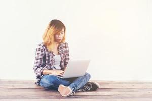 Young hipster woman sitting on wooden floor with crossed legs and using laptop on white background photo