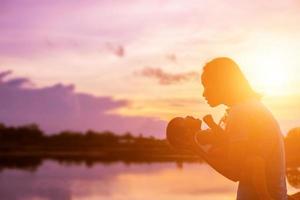 una silueta de una niña feliz los brazos de su madre amorosa para un abrazo, frente a la puesta de sol en el cielo en un día de verano. foto