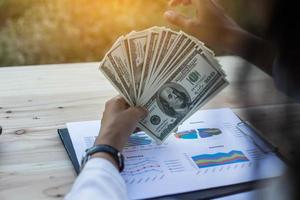 mujer de negocios agarrando los billetes de cien dólares de los Estados Unidos en la mano y algunos puestos en la mesa llenos de gráficos de negocios y cuadernos foto