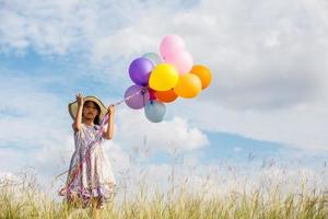 linda niña sosteniendo globos coloridos en el prado contra el cielo azul y las nubes, extendiendo las manos.