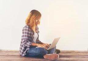 Young hipster woman sitting on wooden floor with crossed legs and using laptop on white background photo