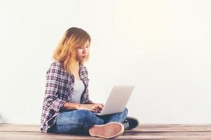 Young hipster woman sitting on wooden floor with crossed legs and using laptop on white background photo
