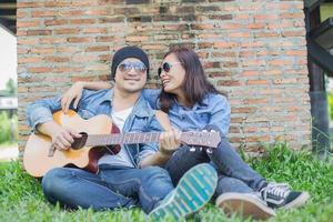 Hipster man playing guitar for his girlfriend outdoor against brick wall, enjoying together. photo