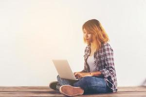 Young hipster woman sitting on wooden floor with crossed legs and using laptop on white background photo