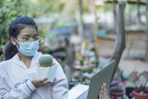 Beautiful Asian girl greenhouse worker selling small plants on social media live stream with mobile. Small tree landscaping and home garden decoration gardener. photo