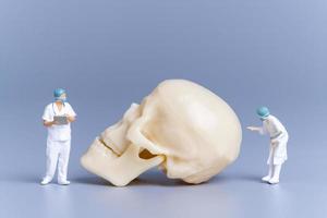 médico de personas en miniatura con un cráneo humano gigante sobre un fondo gris foto