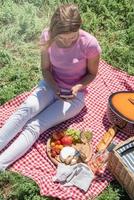 woman in white pants outside having picnic and using smartphone taking photo