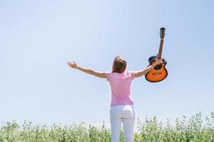 Young woman standing in grass fiel raising her hand to the sky, holding guitar photo
